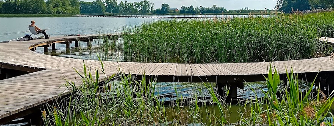 Na zdjęciu widać jezioro i drewniany pomost w kształcie okręgu. Na pomoście po lewej stronie siedzi na leżaku wędkarz. Brzeg jeziora porasta trzcina.